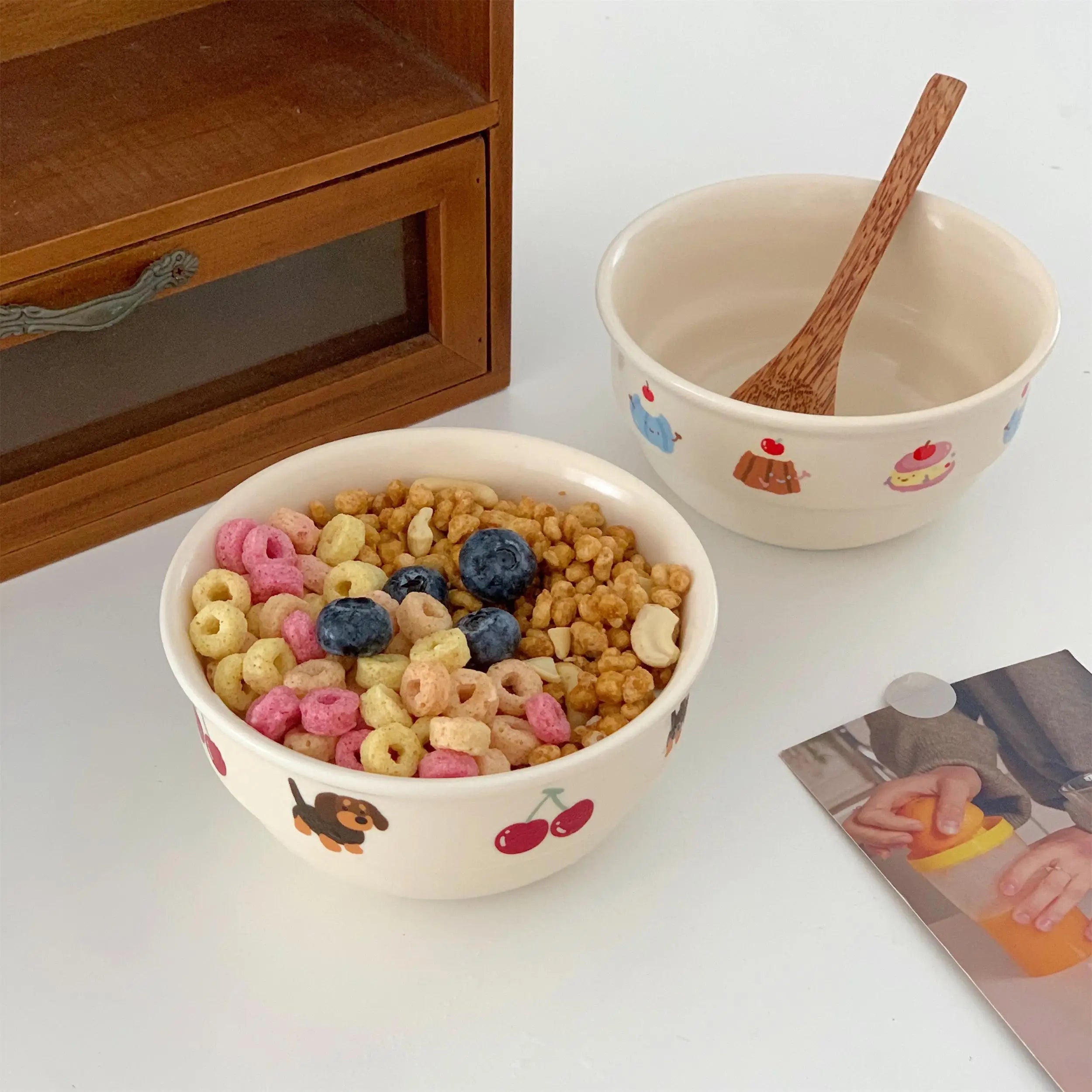 Two kawaii ramen bowls with cereal and a wooden spoon on a white surface, with a wooden cabinet in the background.
