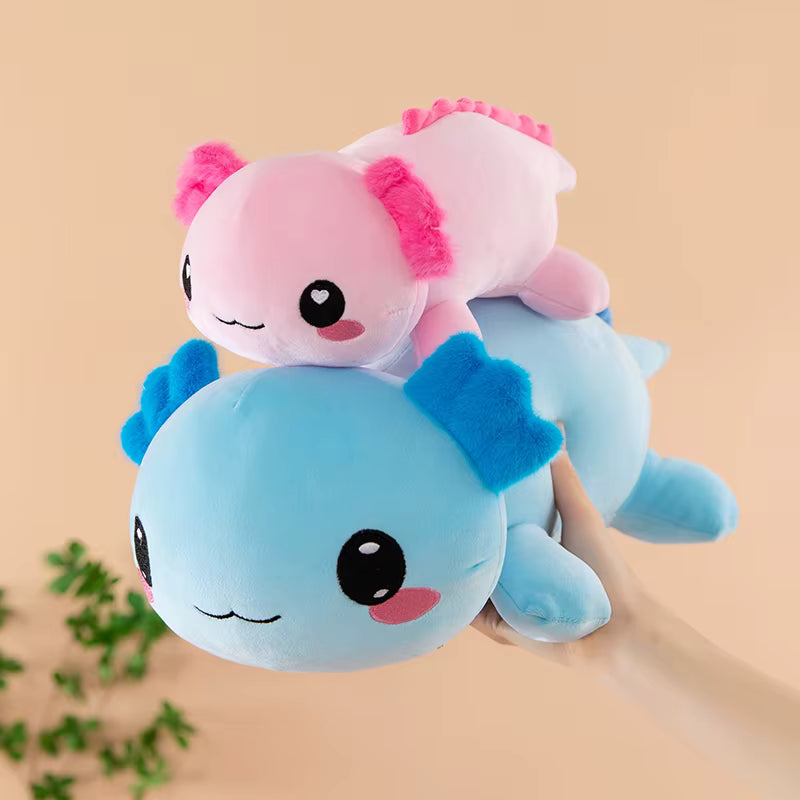 Two cute plush axolotl toys stacked on top of each other, held by a hand against a soft beige background with a small green plant in the corner.