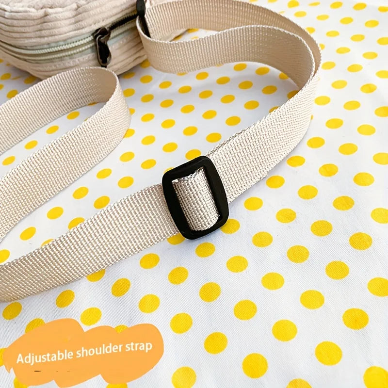 A close-up of a beige adjustable woven shoulder strap with a black plastic buckle, laid on a white fabric background with yellow polka dots. Part of a matching zippered bag is visible in the upper left, and an orange label reads 'Adjustable shoulder strap'.