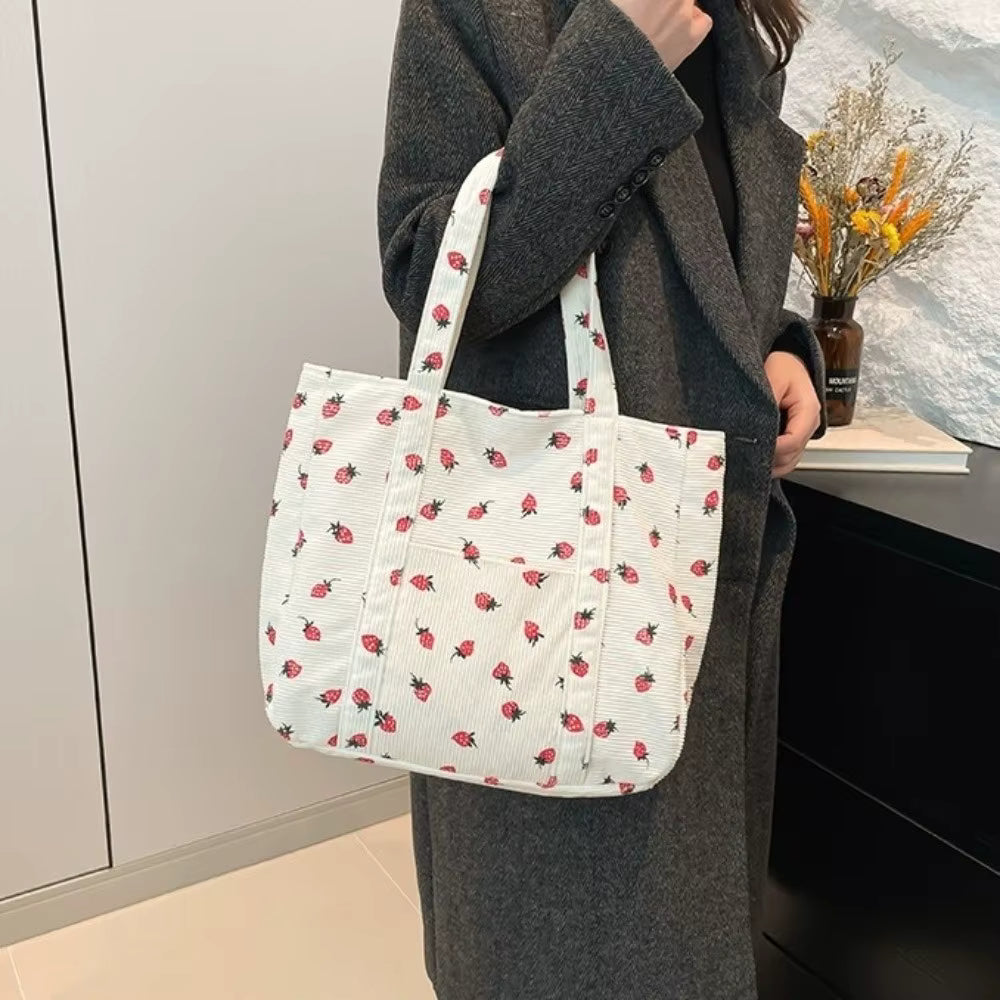 A person wearing a dark gray coat carries a white canvas tote bag decorated with a small red strawberry pattern in an indoor setting.