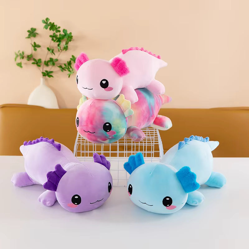 Four cute axolotl plush toys in pastel colors arranged on a white table, with one pink plush and one tie-dye plush displayed on a small wire stand and two more standing on the table in front.
