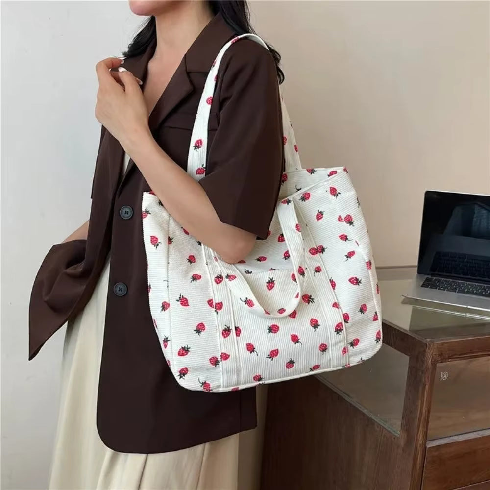 A woman wearing a brown blazer and cream dress carries a large white tote bag with a red strawberry print in an indoor setting.