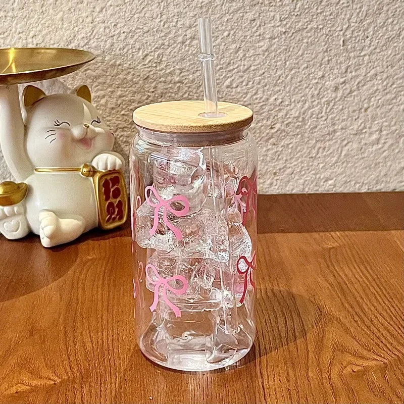 Clear glass kawaii tumbler with wooden lid and straw on a wooden surface, next to a decorative cat figurine.