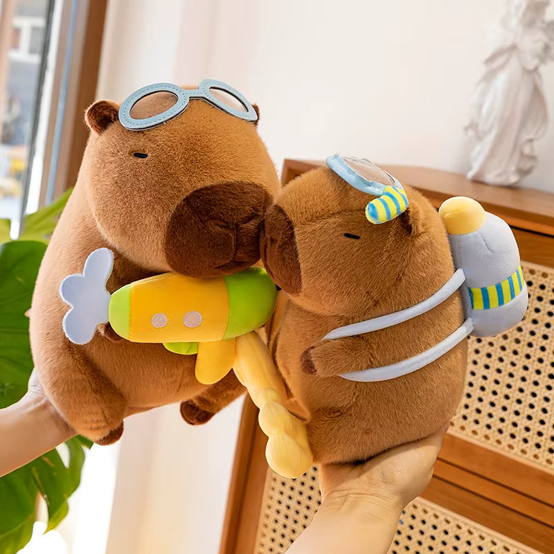 Two brown capybara plush toys being held by hands in a bright indoor setting. One capybara wears light blue goggles and holds a yellow and green toy submarine, while the other wears a blue-and-yellow swim cap and a white backpack with green stripes.