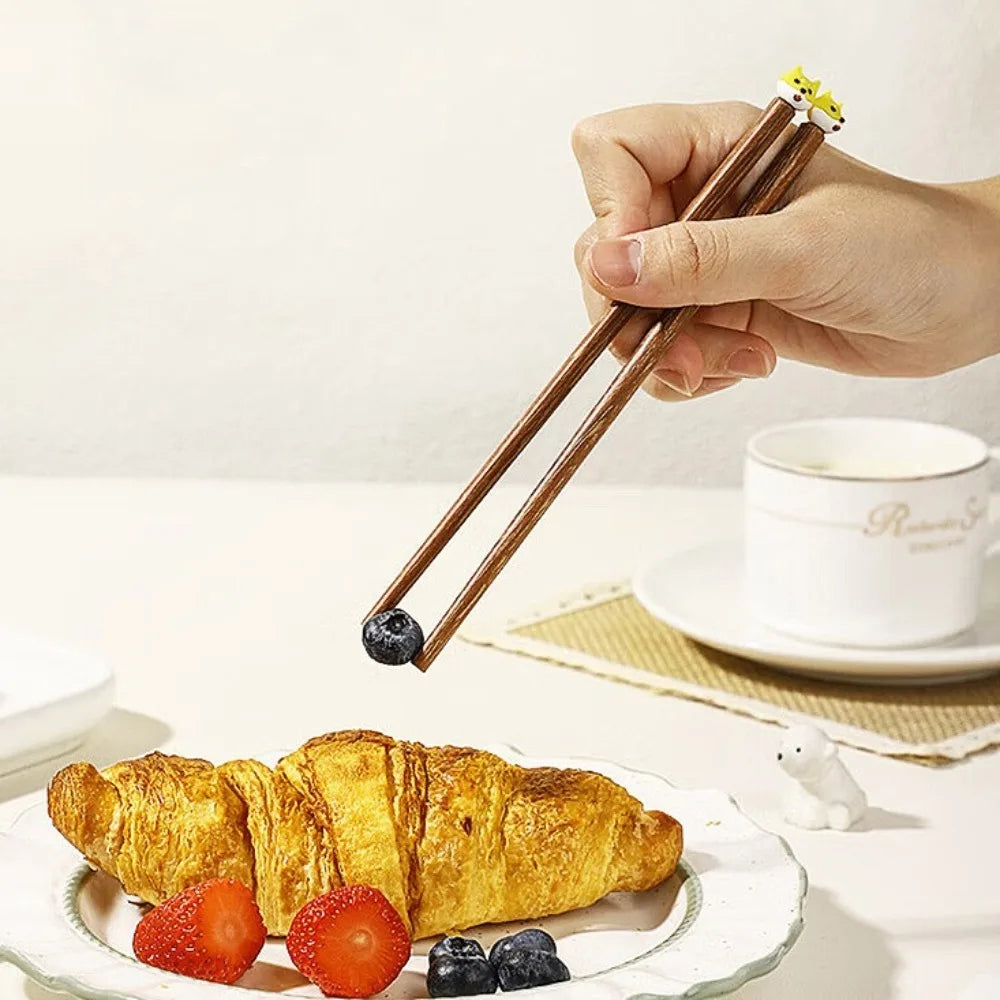 Hand using kawaii chopsticks to pick up a blueberry over a plate with a croissant and berries on a white background.