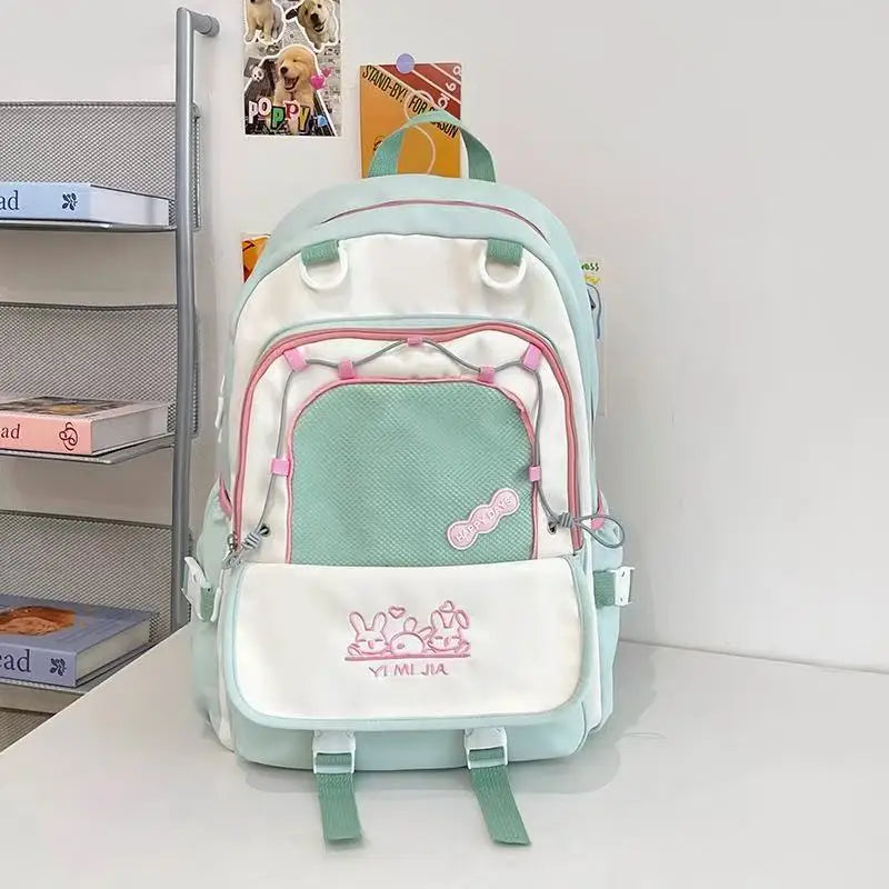A pastel green, pink and white school backpack with pink zipper accents and cartoon bunny graphics on the front pocket, displayed on a white table against a white wall with decorative books and cards.