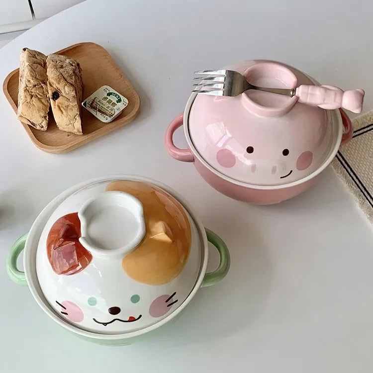 Two cartoon-style ceramic kawaii ramen bowls with lids on a white surface, one pink and one green, with a small wooden tray of bread and butter in the background.