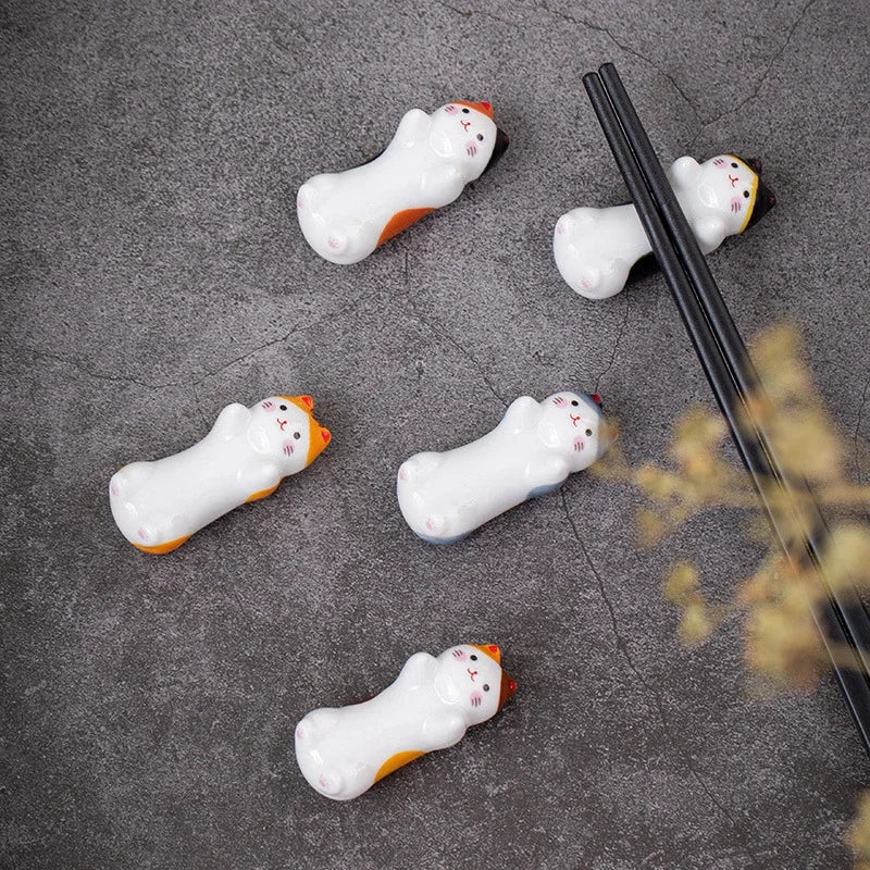 Five cat-shaped ceramic kawaii chopstick holders on a gray surface with a pair of black chopsticks.