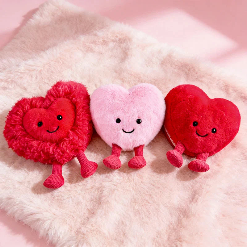 Three heart-shaped kawaii plush toys with smiling faces on a soft pink surface