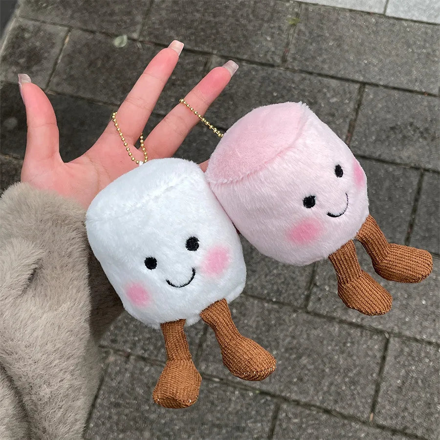 Two kawaii plush toys resembling marshmallows with faces held by a hand on a pavement background.