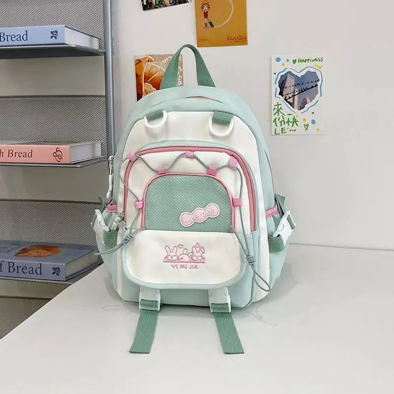 A pastel green, pink and white school backpack with pink zipper accents and cartoon bunny graphics on the front pocket, displayed on a white table against a white wall with decorative books and cards.
