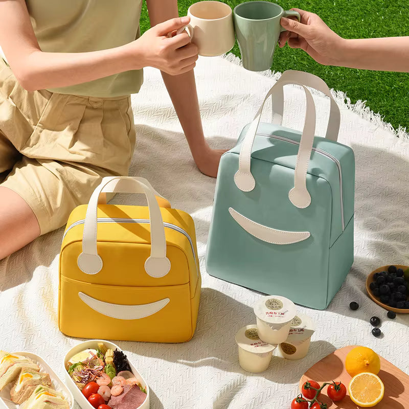 Two people enjoying a picnic with smiley face lunch boxes and food on a blanket outdoors.