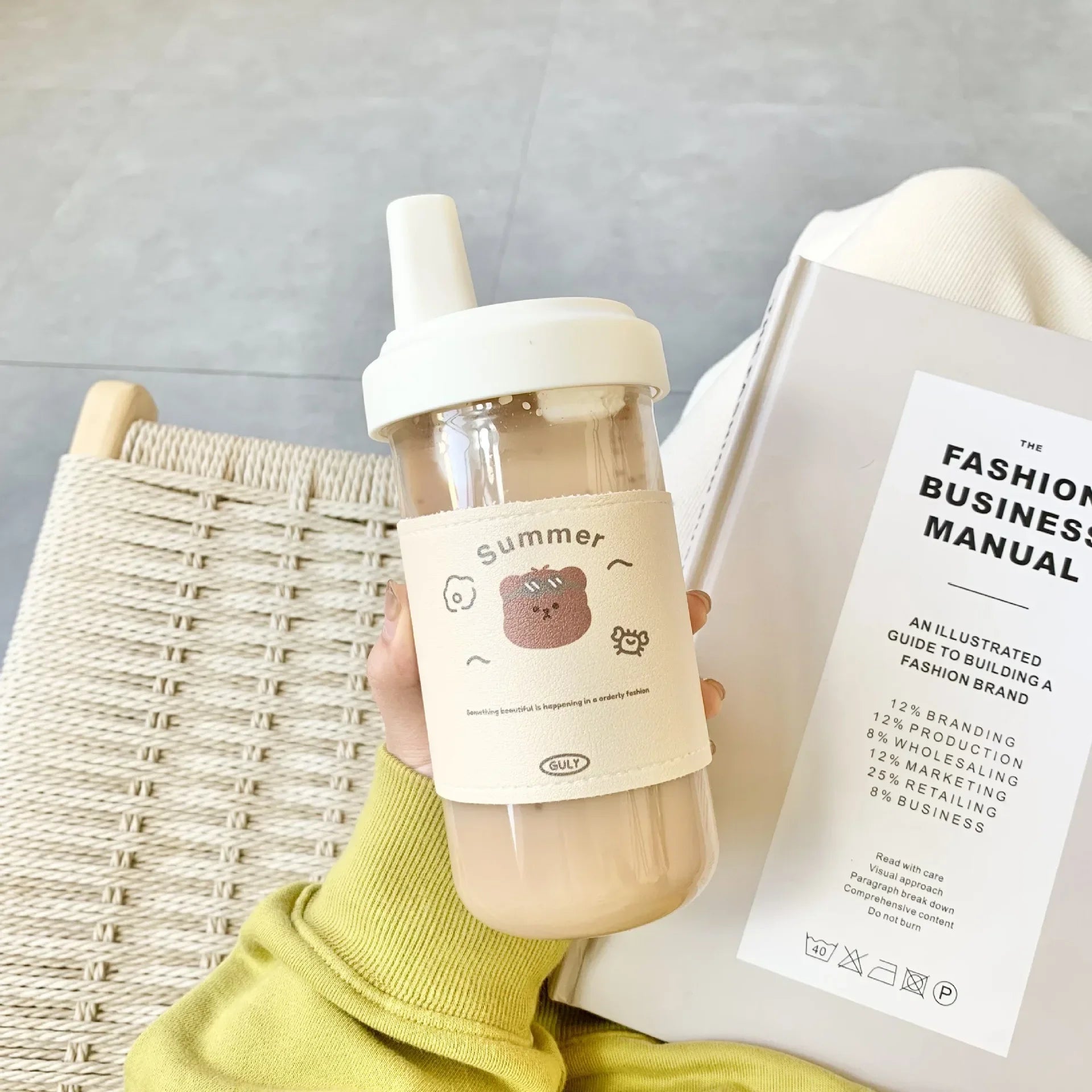 Person holding a kawaii water bottle with a straw and a book titled 'The Fashion Business Manual' on a light gray background.