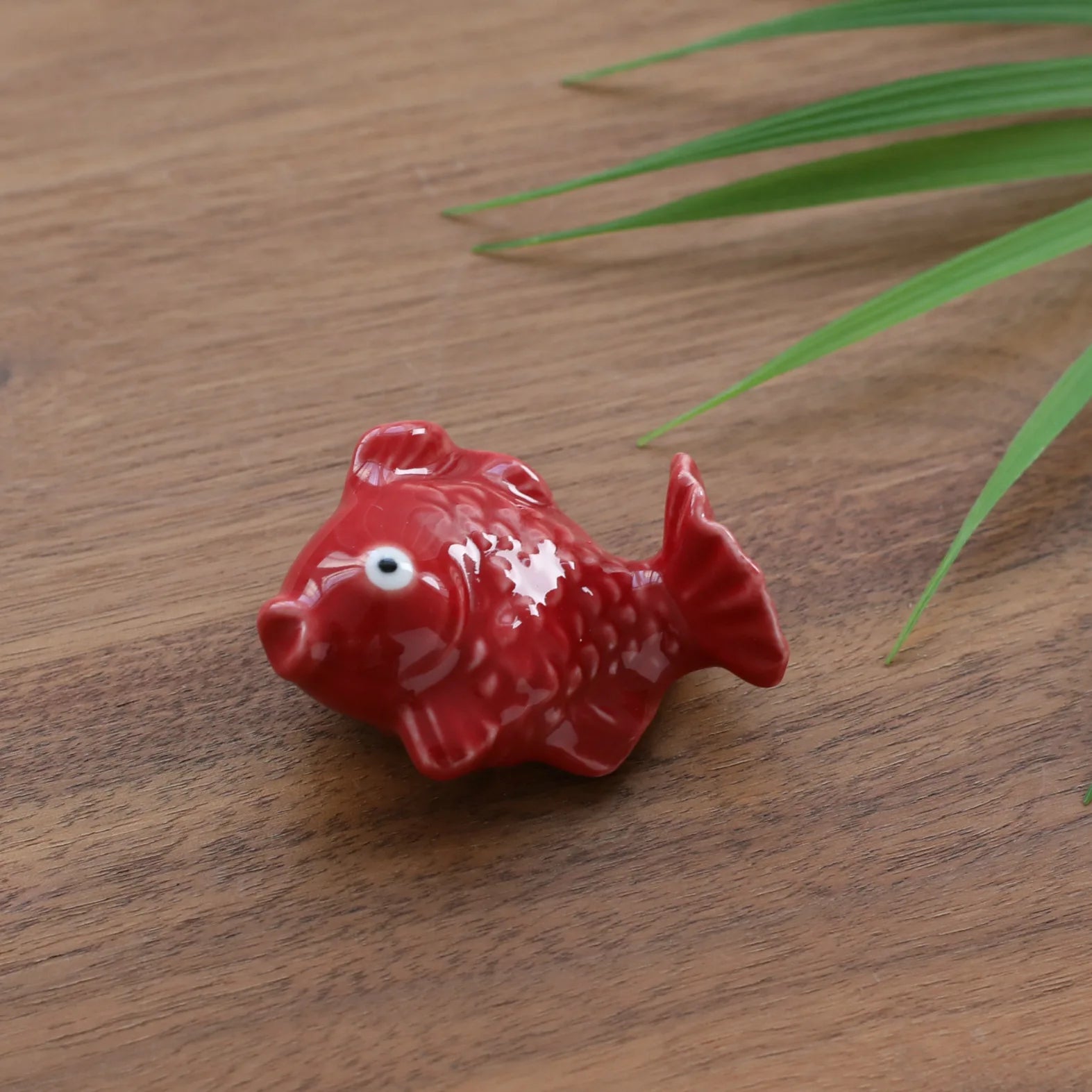 Red ceramic fish kawaii chopstick holders on a wooden surface with green leaves.