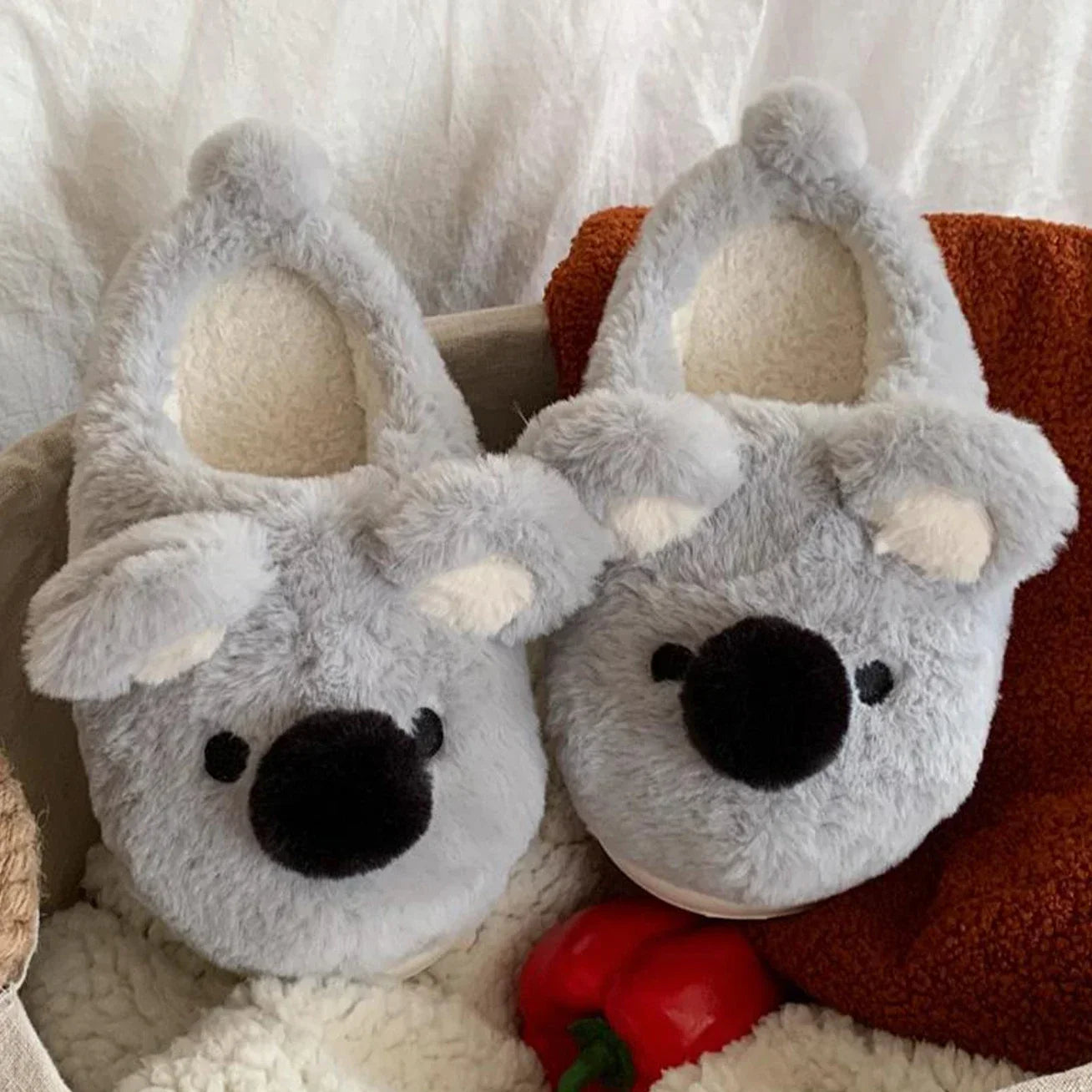 Pair of gray koala kawaii slippers on a white surface with a red toy in the background.