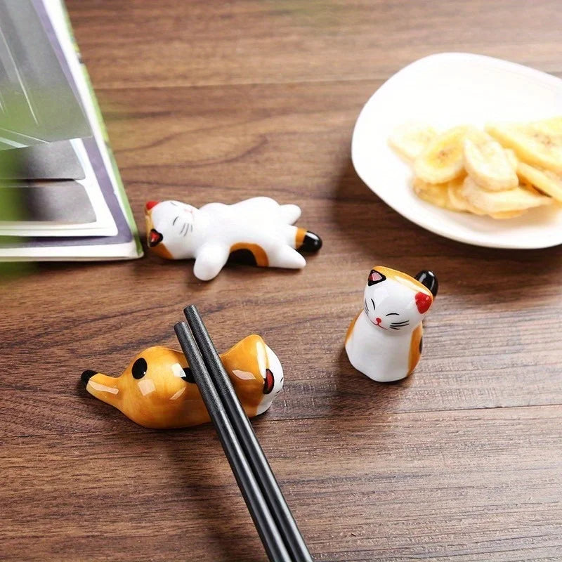 Three cat-shaped kawaii chopstick holder on a wooden surface with a plate of snacks in the background.