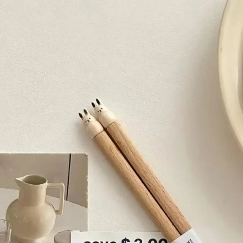 Two wooden kawaii chopsticks with black ends on a white surface, next to a ceramic pitcher.